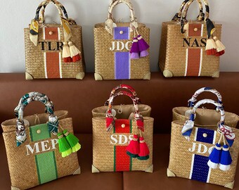 May include: Six woven straw tote bags with rectangular shapes, each featuring a vertical colored stripe, a central monogram, and a patterned silk scarf wrapped around the handles. Each bag includes a pair of decorative tassels attached to the handle.