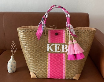 May include: A woven straw tote bag with pink and gold stripes, a white monogram "KEB", and pink tassels. The bag has a pink and white patterned fabric handle.