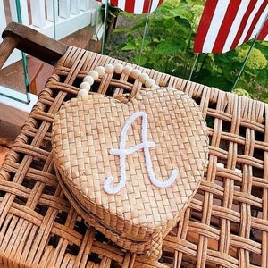 May include: A heart-shaped woven straw bag with a white beaded handle and the letter "A" embroidered on the front. The bag is sitting on a wicker chair with American flags hanging in the background.