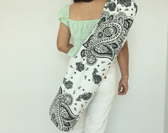May include: A white yoga mat bag with a black paisley pattern. The bag is worn over the shoulder of a person wearing a green and white gingham shirt and white pants.
