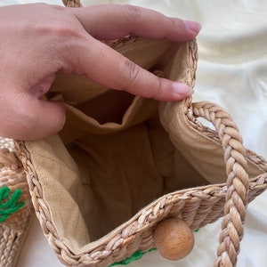 May include: A woven straw bag with a brown rope handle and a green embroidered design. The bag has a beige fabric lining and a wooden toggle closure.