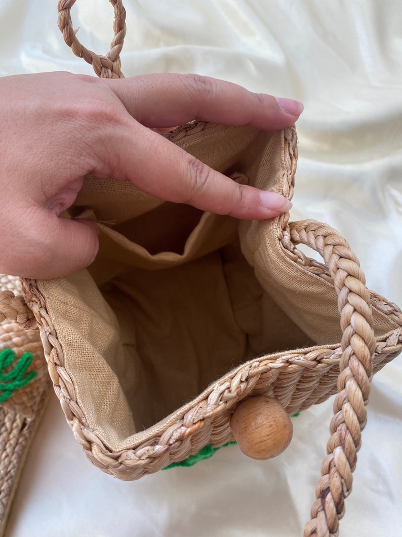 May include: A woven straw bag with a brown rope handle and a green embroidered design. The bag has a beige fabric lining and a wooden toggle closure.