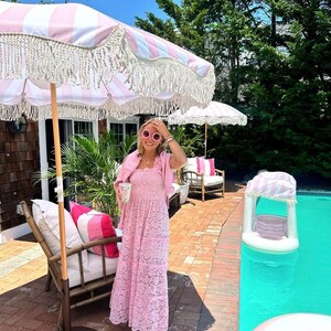 May include: A woman wearing a pink lace dress and pink heart-shaped sunglasses stands by a pool. She is holding a glass of something and is smiling. There are pink and white striped umbrellas in the background.
