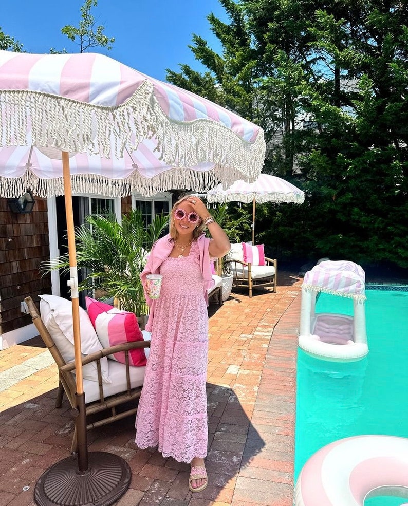May include: A woman wearing a pink lace dress and pink heart-shaped sunglasses stands by a pool. She is holding a glass of something and is smiling. There are pink and white striped umbrellas in the background.