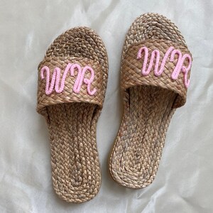May include: A pair of woven straw slides with pink embroidered letters that spell "WWR" on each slide.