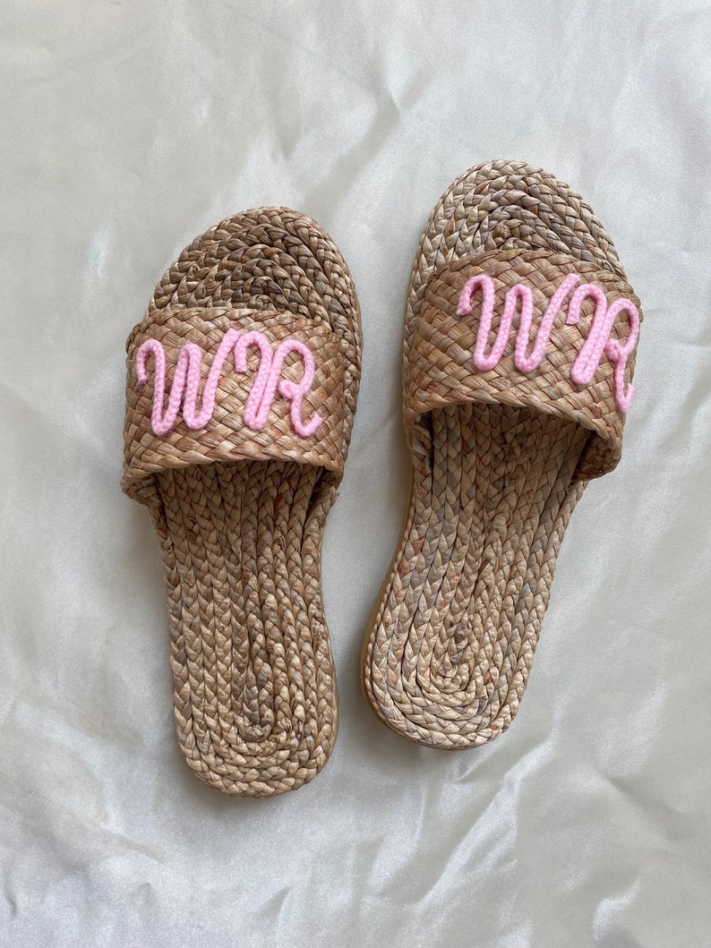 May include: A pair of woven straw slides with pink embroidered letters that spell "WWR" on each slide.