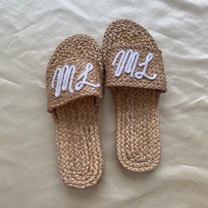 May include: A pair of woven raffia slides with white embroidered initials "ML" on the top. The sandals are light brown and have a textured appearance. The background is a light beige fabric.