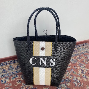 May include: Black woven tote bag with rope handles. The bag features a beige and white stripe design with the embroidered letters "CNS". A wooden button secures the top.