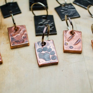 May include: A collection of copper and black metal keychains with paw print designs. The keychains are on a wooden surface.