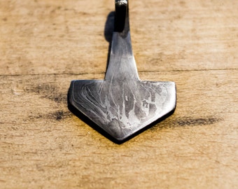 Puede incluir: Primer plano de un colgante de metal con forma de martillo de Thor, Mjolnir. El colgante tiene una superficie texturizada y estampada de color gris oscuro. El mango tiene un tono azul. El colgante está sobre un fondo de madera.