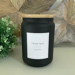 Peut inclure: Pot en verre noir mat avec un couvercle en bambou. Le pot a une étiquette blanche avec le texte "YOUR TEXT" dessus.
