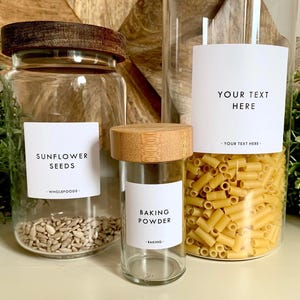 Peut inclure: Trois bocaux en verre transparent avec des couvercles en bois et des étiquettes blanches. Les bocaux contiennent des graines de tournesol, de la levure chimique et des pâtes. Les étiquettes indiquent "GRAINES DE TOURNESOL", "LEVURE CHIMIQUE" et "VOTRE TEXTE ICI".