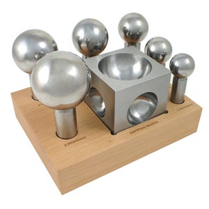 Puede incluir: Un juego de seis punzones de bola de metal y un bloque de dapping, todos dispuestos en un soporte de madera. Los punzones son de diferentes tamaños y están etiquetados con su diámetro en pulgadas y milímetros. El bloque de dapping tiene una hendidura redonda en el centro.