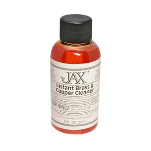 May include: A small, clear bottle with a red liquid inside. The bottle is labeled "JAX Instant Brass & Copper Cleaner" and has a warning label. The bottle is 2 fluid ounces or 59 milliliters.