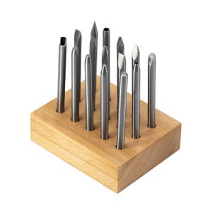 May include: A set of twelve metal punches with various tip shapes, including pointed, flat, and hollow ends, arranged upright in a rectangular light-coloured wooden block with twelve drilled holes to hold each tool securely in place.