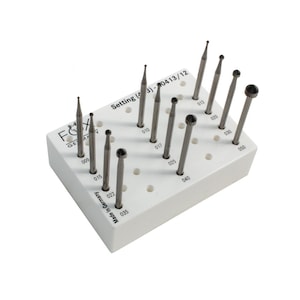 May include: A white rectangular holder containing various metal drill bits. The holder is marked with "FX GERMANY" and "Setting (3) - 0413/12". The drill bits are of different shapes and sizes, arranged in a grid pattern. Made in Germany.