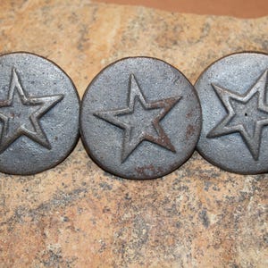May include: Three weathered, circular metal knobs with a raised star design. The knobs are a dark gray color with a slightly textured surface, and the star has a rustic, aged appearance. These decorative knobs could be used for furniture or other craft projects.