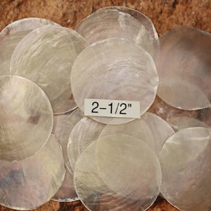 May include: A collection of iridescent, round, mother-of-pearl shell pieces. Each shell is approximately 2-1/2 inches in diameter. The shells have a pearlescent sheen and are arranged on a textured, brown surface.