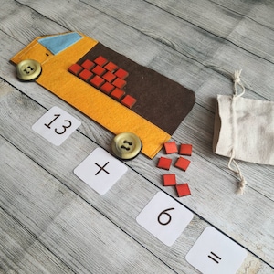 Può includere: Un camion giocattolo di feltro con un corpo marrone e giallo, che trasporta blocchi rossi. Il camion viene utilizzato per insegnare la matematica, con carte che mostrano l'equazione 13 + 6 = .