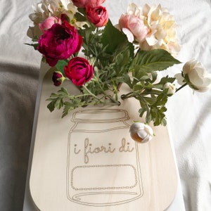 Può includere: Un cartello in legno a forma di vaso, con le parole "i fiori di" e un'etichetta vuota. Il cartello è decorato con una varietà di fiori artificiali colorati, tra cui fiori rosa, rossi e bianchi, e foglie verdi.