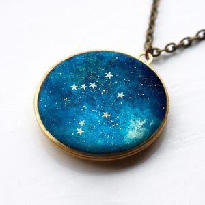 Puede incluir: Un collar con un colgante redondo de vidrio azul y blanco que presenta una constelación de estrellas. El colgante está engastado en un marco dorado.