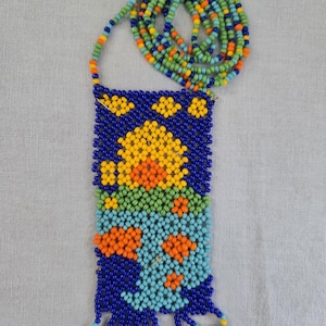 May include: A beaded necklace with a rectangular pendant. The pendant is primarily blue, with a floral design in yellow, orange, and turquoise beads. The necklace cord is a mix of green, blue, orange, and yellow beads.