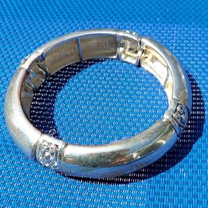 May include: Silver-toned bracelet with a rounded design. The bracelet has decorative elements with a shield-like shape. The bracelet is on a blue surface.