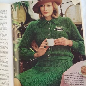 May include: A green knitted jacket and skirt set with a brown hat. The jacket has a button closure and a small brooch. The skirt is slightly flared. The text "Trim battle jacket, precisely buttoned in silver, just grazes the top of a slightly flared skirt. Suit is worked in alternating panels of plain afghan stitch crochet and purl afghan stitch. Directions on opposite page." is visible in the image.