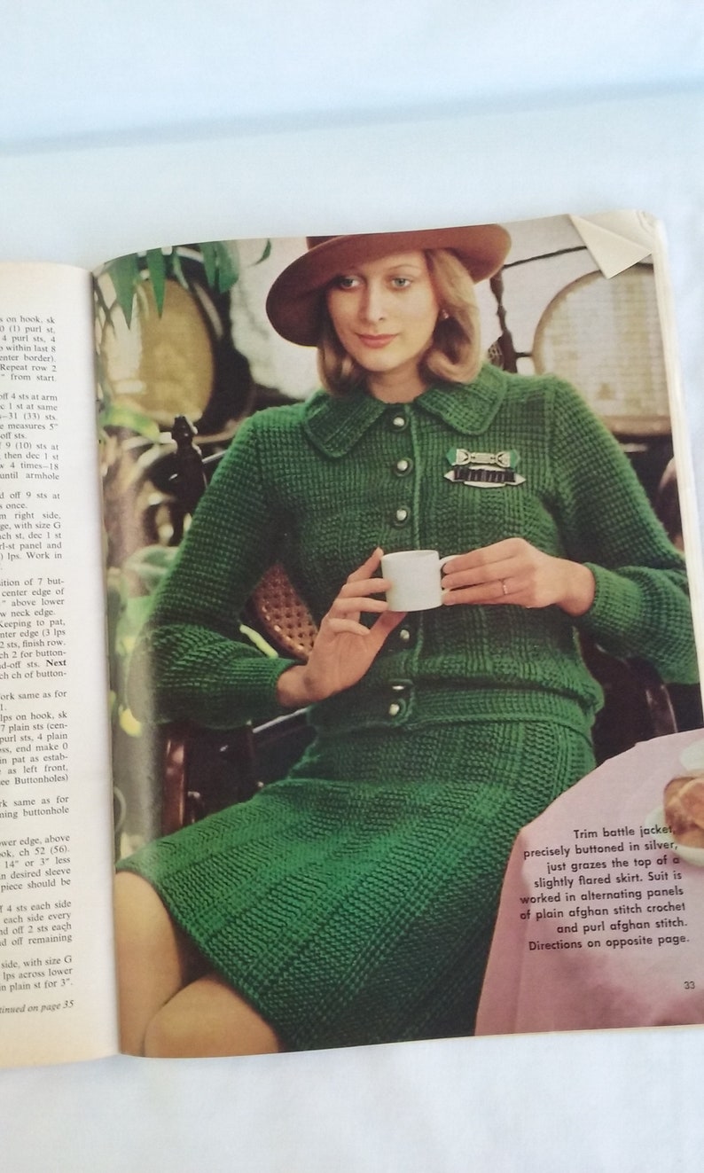 May include: A green knitted jacket and skirt set with a brown hat. The jacket has a button closure and a small brooch. The skirt is slightly flared. The text "Trim battle jacket, precisely buttoned in silver, just grazes the top of a slightly flared skirt. Suit is worked in alternating panels of plain afghan stitch crochet and purl afghan stitch. Directions on opposite page." is visible in the image.