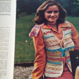 May include: A crochet vest with a colorful striped pattern and a tie closure. The vest is worn over a long-sleeved orange shirt and a red skirt. The image also shows a close-up of a girl wearing a similar vest.