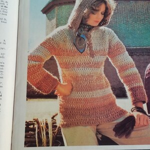 May include: A hooded sweater with a tweed stripe pattern in shades of brown and beige. The sweater is crocheted and has a long, loose fit. The hood is attached and has a long, flowing shape. The sweater is worn by a model with long, brown hair. The model is standing in front of a stone wall.