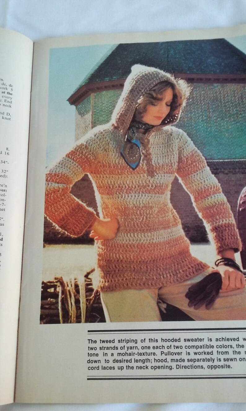 May include: A hooded sweater with a tweed stripe pattern in shades of brown and beige. The sweater is crocheted and has a long, loose fit. The hood is attached and has a long, flowing shape. The sweater is worn by a model with long, brown hair. The model is standing in front of a stone wall.