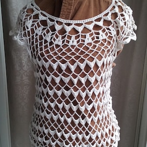 May include: White crochet top with a delicate, lacy pattern. The top has a scoop neck and short sleeves.