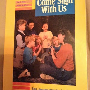 May include: A book titled "Come Sign With Us" with a group of children signing. The book cover features a yellow background with red and blue stripes. The book is about sign language activities for children.