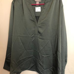 May include: A long-sleeved, olive green blouse with a V-neck and a slight gathering detail at the neckline. The blouse is made of a smooth, silky fabric and hangs on a pink hanger. The tag shows the brand logo and price.