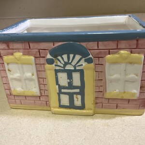 May include: A rectangular ceramic planter shaped like a house. The planter features a brick pattern in shades of pink and red, with a blue roofline. It has a yellow door and window frames, with white windows. The planter is designed for decorative use.