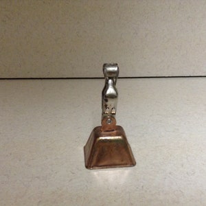 May include: A small, silver-colored bell with a copper-colored base. The bell is attached to a silver-colored handle.