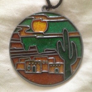 May include: A round, metal ornament with a stained glass design depicting a desert scene. The scene includes a setting sun, a cactus, and a row of adobe houses. The ornament is decorated with glitter.