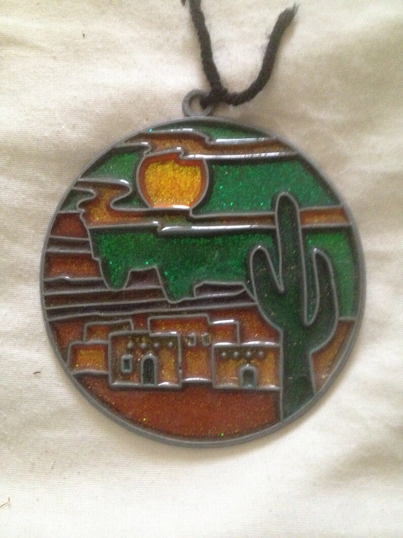 May include: A round, metal ornament with a stained glass design depicting a desert scene. The scene includes a setting sun, a cactus, and a row of adobe houses. The ornament is decorated with glitter.