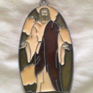 May include: A stained glass window hanging ornament featuring a depiction of Jesus Christ in a brown robe with a light brown background and green grass at the bottom.