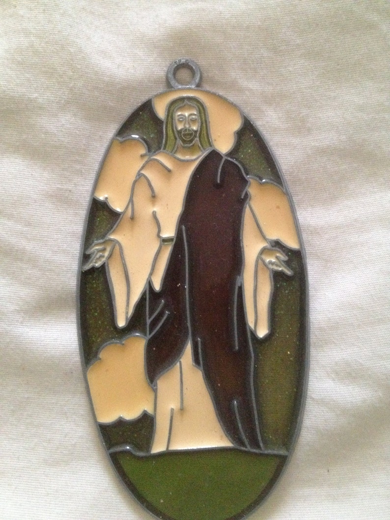 May include: A stained glass window hanging ornament featuring a depiction of Jesus Christ in a brown robe with a light brown background and green grass at the bottom.