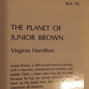 May include: A book cover with the title "The Planet of Junior Brown" by Virginia Hamilton. The cover is a light brown color with a gold background. The text is in black.