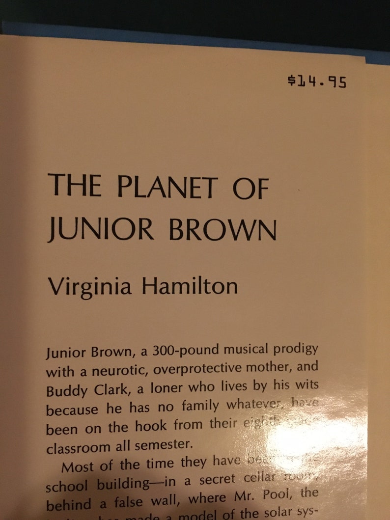 May include: A book cover with the title "The Planet of Junior Brown" by Virginia Hamilton. The cover is a light brown color with a gold background. The text is in black.