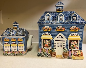 May include: A blue ceramic teapot and sugar bowl, both in the shape of houses. The teapot has a spout and handle, while the sugar bowl is a larger, house-shaped container. Both feature floral accents and detailed painting.