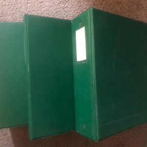 May include: Three dark green binders. The binders are arranged side by side, with the spine of the rightmost binder facing the viewer. The binders have a rectangular shape and a smooth surface. The rightmost binder has a white label holder.