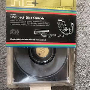 May include: A new Compact Disc Cleaner kit in a clear plastic case. The packaging is black, yellow, and green with the text "HE+" and "Compact Disc Cleaner". The kit includes a CD cleaner and a CD spray bottle. The product is designed to remove oils, fingerprints, and soil from compact discs.