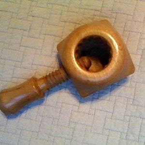 May include: A wooden nutcracker with a square head and a round opening. The handle is attached to a threaded screw mechanism. The nutcracker is made of light-colored wood and is designed for cracking nuts.