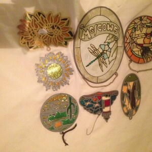 May include: Seven stained glass ornaments with various designs, including a dragonfly, a lighthouse, a sunflower, and a floral pattern. The ornaments are all hanging from chains.