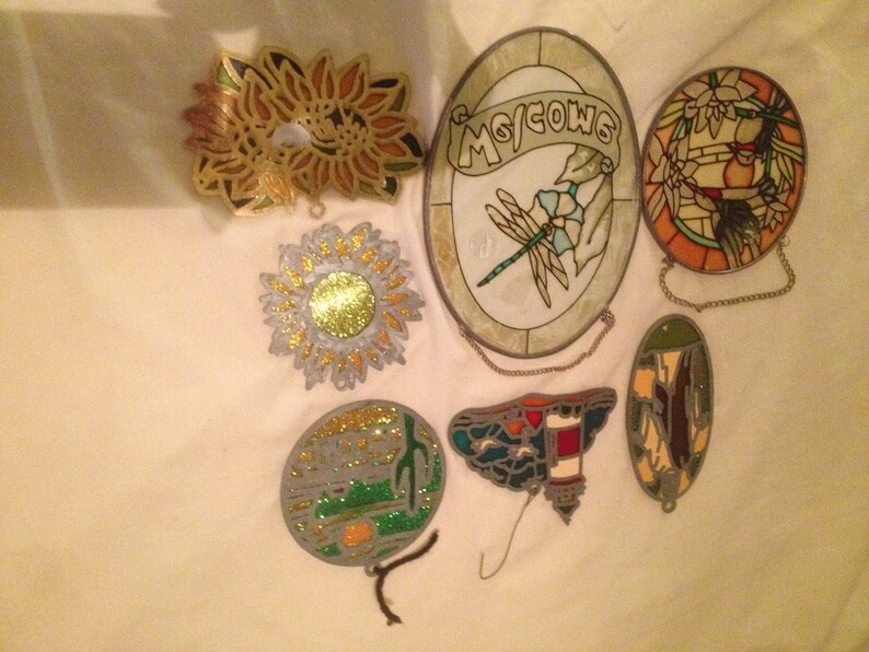 May include: Seven stained glass ornaments with various designs, including a dragonfly, a lighthouse, a sunflower, and a floral pattern. The ornaments are all hanging from chains.