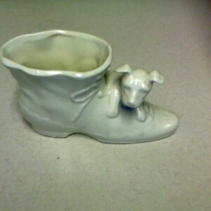 Puede incluir: Macetero de cerámica blanca con forma de zapato con una pequeña figura de perro en la parte superior.